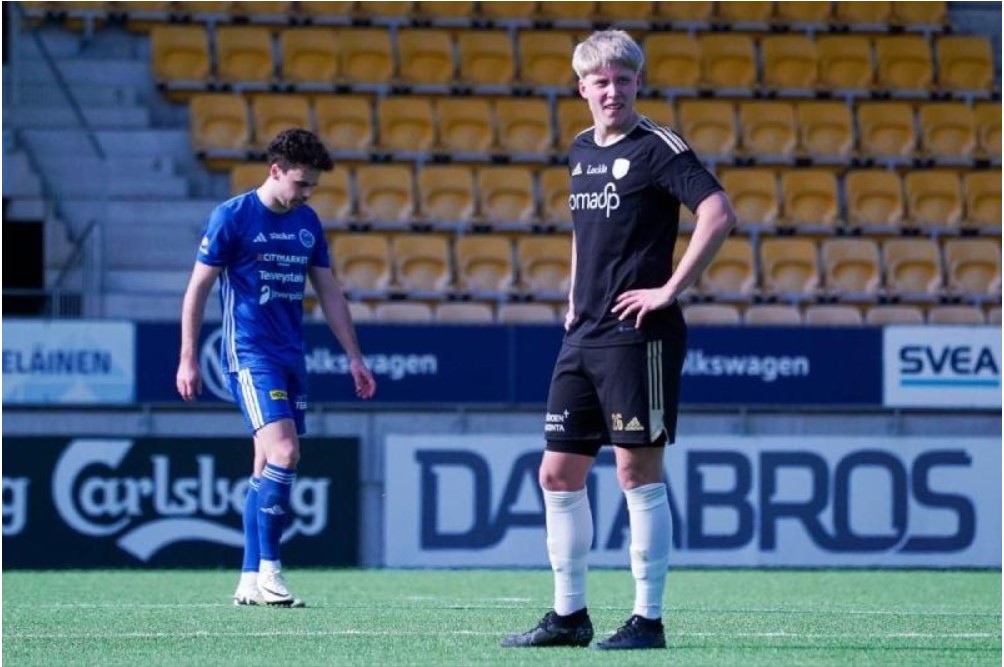 A person in a black shirt and white socks standing on a green field with a person in a blue uniformDescription automatically generated